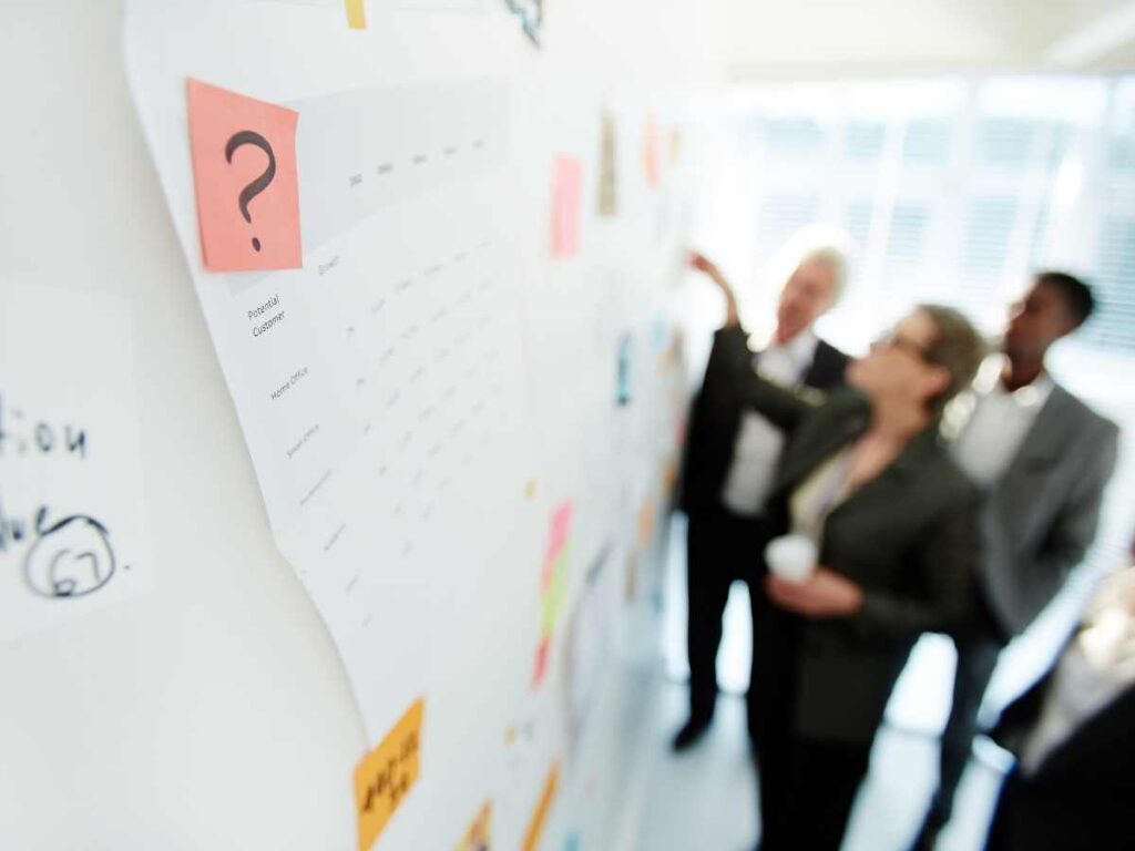 Grupo de profissionais discutindo e apontando para gráficos em uma parede, representando o processo colaborativo e reflexivo ao concluir um projeto acadêmico.