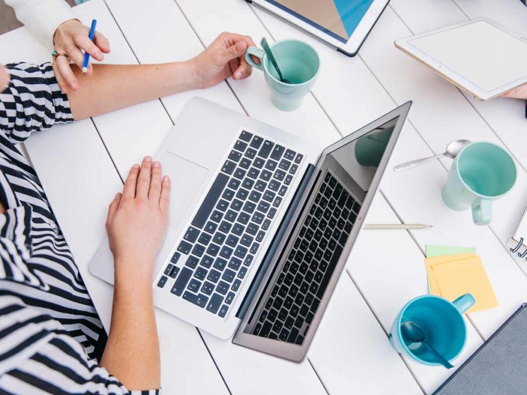 Mãos de estudantes ao redor de um laptop, trabalhando e discutindo, com materiais de escritório coloridos, representando a criação de uma tese.