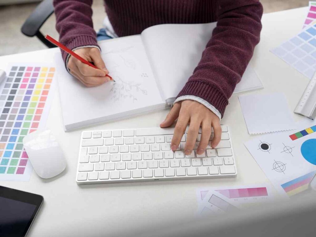 Pessoa desenhando no caderno enquanto utiliza uma paleta de cores para um projeto de design gráfico.