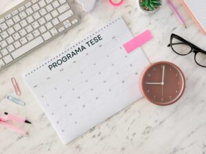 Mesa de trabalho com calendário de programa de tese, relógio e materiais de escritório simbolizando organização e gestão do tempo.