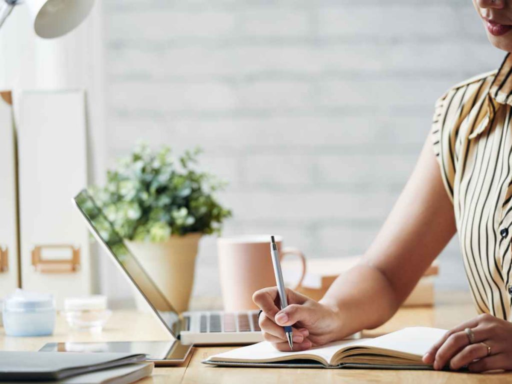 Pessoa escrevendo em caderno com laptop e planta ao fundo